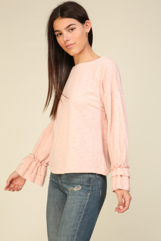 Woman wearing a light pink ruffle cuff long sleeve top with relaxed fit paired with distressed blue jeans against a beige background
