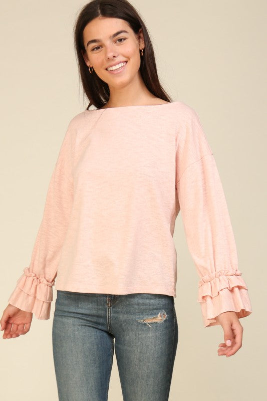 Woman wearing a light pink relaxed fit top with ruffle layered cuffs and boat neckline paired with ripped blue jeans