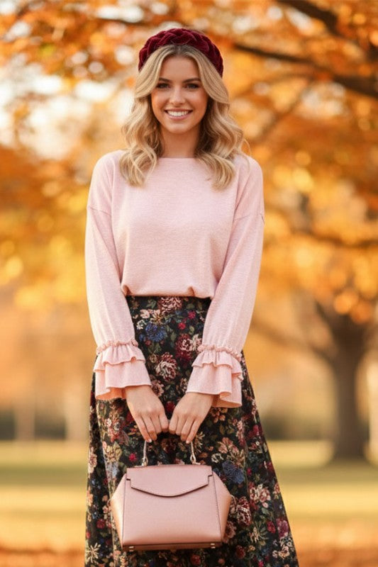 Woman wearing a light pink long sleeve top with layered ruffle cuffs paired with a floral skirt and matching handbag outdoors