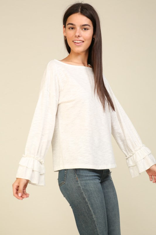 Woman wearing an off-white long sleeve top with layered ruffle cuffs and boat neckline paired with blue jeans