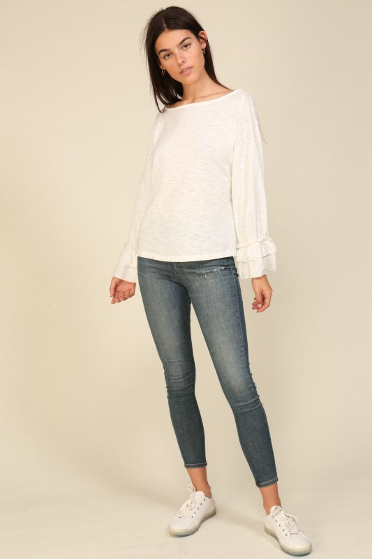 Woman wearing an off-white relaxed fit long sleeve top with layered ruffle cuffs and a boat neckline paired with blue slim jeans