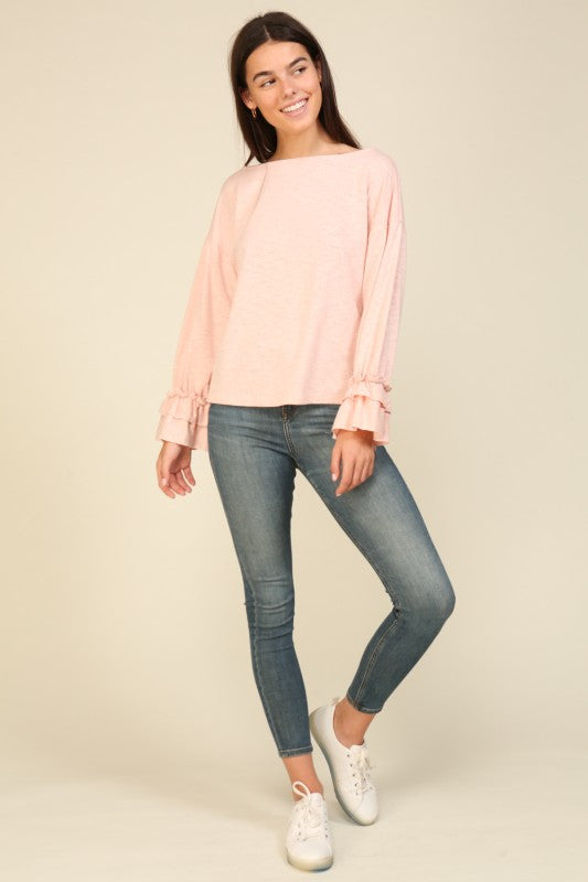 Woman wearing a light pink relaxed fit long sleeve top with layered ruffle cuffs paired with slim blue jeans and white sneakers
