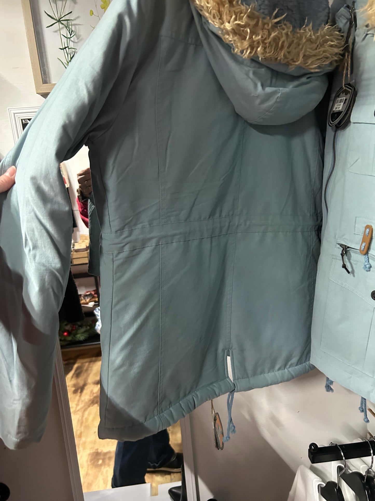 Light green STS Ranchwear coat with faux fur hood trim and adjustable drawstrings shown from the back inside a store